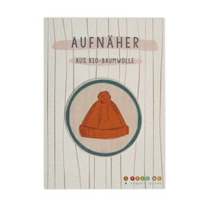 Könnte beinhalten: Eine runde, orangefarbene Mütze mit einem weißen Bommel oben. Die Mütze befindet sich auf einem weißen Hintergrund mit dem Text "AUFNÄHER AUS, BIO-BAUMWOLLE" und "TELL ME organic goods".
