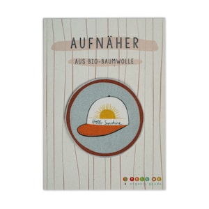 Könnte beinhalten: Ein runder, weißer und orangefarbener Bügelaufkleber mit einer Cartoon-Illustration einer Baseballkappe mit einer Sonne und dem Text "Hello Sunshine". Der Patch befindet sich auf einer Karte mit dem Text "AUFNÄHER AUS BIO-BAUMWOLLE" und dem Logo "TELL ME organic goods".