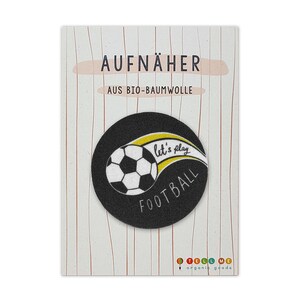 Op de afbeelding: Zwart-witte voetbalpatch met gele tekst die "Let's play football" zegt op een witte achtergrond. De patch bevindt zich op een witte kaart met de tekst "Aufnäher" en "Aus Bio-Baumwolle" in zwart.