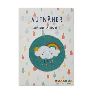 Può includere: Una toppa rotonda, color verde acqua, con un design a nuvola sorridente, arcobaleno e gocce di pioggia. La toppa è su una carta con il testo tedesco "AUFNÄHER AUS BIO-BAUMWOLLE" e un motivo di gocce di pioggia colorate. Il logo del marchio "TELL ME organic goods" è in basso.
