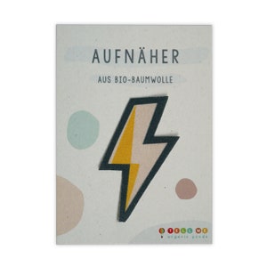 Può includere: Un toppa a forma di fulmine giallo e bianco da stirare con un contorno verde scuro. La toppa è realizzata in cotone biologico ed è etichettata "AUFNÄHER AUS BIO-BAUMWOLLE" e "TELL ME organic goods".