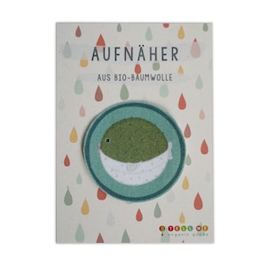 May include: A green and white pufferfish iron-on patch on a white card with the text "AUFNÄHER AUS BIO-BAUMWOLLE" and a colourful "TELL ME organic goods" logo.