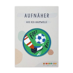 Könnte beinhalten: Ein grün-blauer Bügelaufkleber mit einer Cartoon-Illustration eines Fußes, der einen Fußball tritt. Der Aufnäher sagt "Foot Ball" in weißer Umrandung. Der Text "Aufnäher aus Bio-Baumwolle" ist auf dem weißen Hintergrund gedruckt.