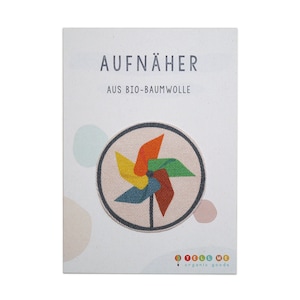 Può includere: Un cartoncino bianco con il testo "AUFNÄHER AUS BIO-BAUMWOLLE" e una toppa a forma di girandola colorata su uno sfondo beige. La girandola è realizzata in tessuto rosso, arancione, giallo, verde e blu. Il cartoncino riporta anche il testo "TELL ME organic goods" in basso.