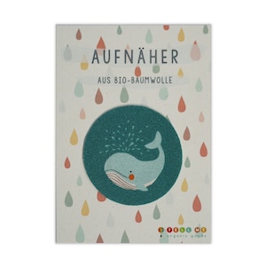 Może przedstawiać: Naszywka z tkaniny w kolorze turkusowym z białym wielorybem, który wydmuchuje wodę. Naszywka jest okrągła i zawiera tekst "AUFNÄHER AUS BIO-BAUMWOLLE" oraz "TELL ME organic goods" na kartce.