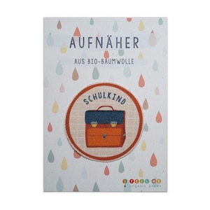 Könnte beinhalten: Ein runder, orangefarbener und weißer Bügelaufkleber mit dem Text "Schulkind" und einem Cartoon-Bild eines roten und blauen Schulranzens. Der Aufnäher ist aus Bio-Baumwolle und trägt die Aufschrift "Aufnäher aus Bio-Baumwolle Tell Me organic goods".