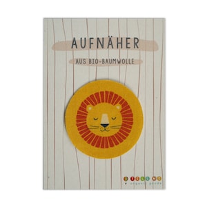Pode incluir: Um patch redondo de leão amarelo com uma juba vermelha e olhos fechados, em um cartão com o texto "AUFNÄHER AUS BIO-BAUMWOLLE". O patch é feito de algodão orgânico e tem o logotipo "TELL ME organic goods".
