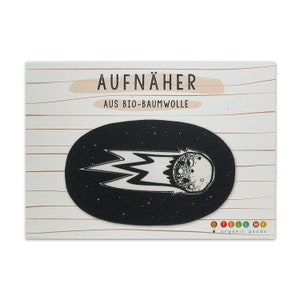 Puede incluir: Un parche termoadhesivo blanco y negro de un cometa con una cola de llamas. El parche tiene forma ovalada y un fondo blanco con detalles negros. El texto "AUFNÄHER AUS BIO-BAUMWOLLE" está impreso en una tarjeta blanca detrás del parche.