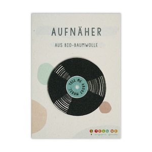 Könnte beinhalten: Ein schwarzer und weißer Vinyl-Platten-Aufnäher mit dem Text "Tell Me About Music" in Türkis auf dem Label. Der Aufnäher befindet sich auf einer weißen Karte mit dem Text "Aufnäher aus Bio-Baumwolle" und "Tell Me organic goods" in bunter Schrift.