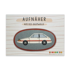 Könnte beinhalten: Weißer Auto-Aufnäher mit rotem Rand auf grauem Hintergrund. Der Aufnäher ist oval und trägt den Schriftzug "AUFNÄHER AUS BIO-BAUMWOLLE" in schwarzer Schrift. Der Text "TELL ME organic goods" befindet sich in der rechten unteren Ecke des Bildes.