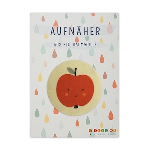 Könnte beinhalten: Ein roter Apfel mit einem lächelnden Gesicht auf einem gelben Stoff-Patch. Der Patch ist mit "AUFNÄHER AUS BIO-BAUMWOLLE" und "TELL ME organic goods" beschriftet.