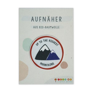 May include: A white, round iron-on patch with a red border. The patch features a black mountain with a white peak and a blue mountain behind it. The text "Up to the Highest Mountains" is printed in black around the mountains.