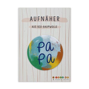 Könnte beinhalten: Ein runder, bunter Bügelaufkleber mit dem Wort "Pa Pa" in blauer Schrift. Der Aufkleber ist aus Bio-Baumwolle und für Kleidung gedacht.