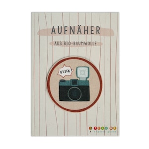 Puede incluir: Un parche redondo de tela marrón con un borde rojo. El parche presenta una ilustración de dibujos animados de una cámara vintage con flash y la palabra "KLICK" en un globo de diálogo. El texto "AUFNÄHER AUS BIO-BAUMWOLLE" está impreso en la tarjeta.