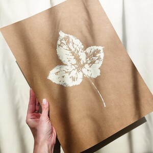 Puede incluir: Una hoja rectangular de papel kraft marrón con una impresión de hoja blanca centrada. El diseño de la hoja muestra detalles intrincados de las venas y un borde dentado, representados en tinta blanca texturizada sobre el papel beige.