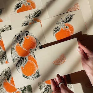 May include: A collection of art prints featuring orange fruit illustrations. The prints showcase oranges with green leaves and floral accents. One print is held by a person, displaying a detailed orange slice.