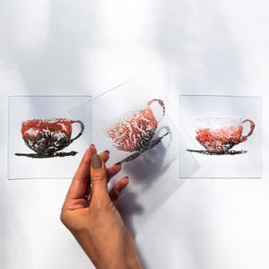 Może przedstawiać: Trzy przezroczyste kwadratowe panele, każdy z unikalnym czerwono-czarnym wzorem filiżanki do herbaty. Filiżanki do herbaty są przedstawione ze skomplikowanymi wzorami, a jeden panel jest trzymany przez dłoń. Filiżanki do herbaty znajdują się na białym tle.