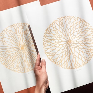 May include: Two art prints featuring gold-colored, circular floral designs on white paper. One print displays a detailed, radiating pattern, while the other showcases a more organic, flowing design. The prints are held by a hand, suggesting a presentation or display.