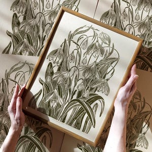 Op de afbeelding: Een ingelijste print van een botanische illustratie met een groep sneeuwklokjes in donkergroen. De print is op een crèmekleurige achtergrond en heeft een houten lijst.