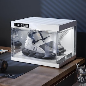 May include: A white and clear shoe display case with a pair of black and white sneakers inside. The case has a white top and a clear front, with the text "ANT BOX" visible. The sneakers have a unique design.