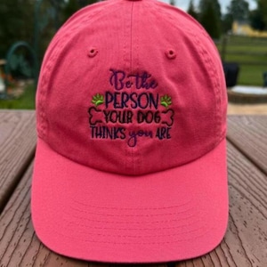Puede incluir: Gorra de béisbol roja con visera curva. La gorra tiene texto bordado que dice "Be the PERSON YOUR DOG thinks you ARE" en morado y verde. El sombrero está hecho de una tela suave.