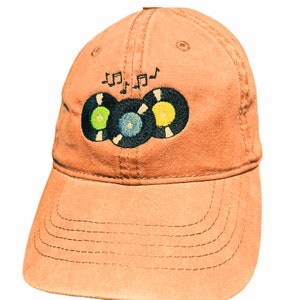 May include: A brown baseball cap with an embroidered design of three vinyl records and musical notes. The records are black with green, blue, and yellow accents.