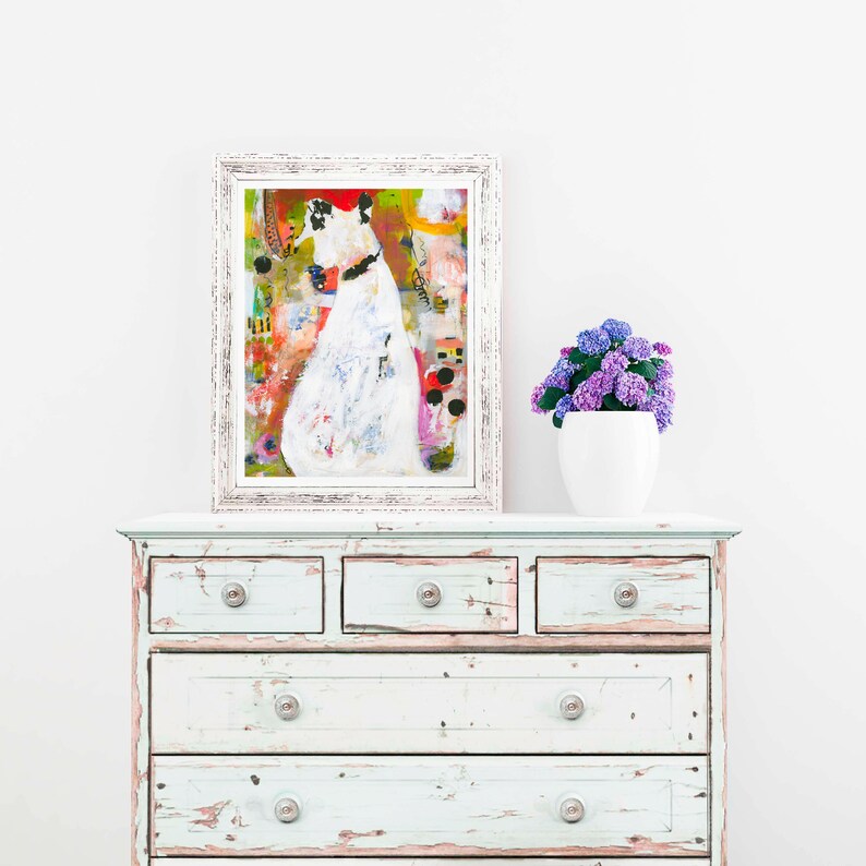 Op de afbeelding: Een witte commode met zes laden en verweerde witte verf. De commode heeft zilveren knoppen. Een ingelijst abstract schilderij van een witte figuur met kleurrijke accenten hangt boven de commode. Een witte plantenbak met paarse bloemen staat op de commode.