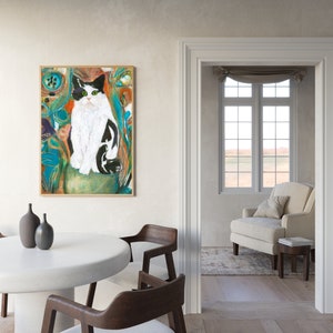 May include: A painting of a black and white cat with green eyes sitting in a colorful, abstract background. The painting is framed in a light wood frame and is hanging on a white wall.