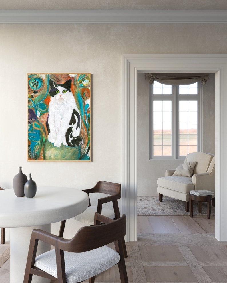 May include: A painting of a black and white cat with green eyes sitting in a colorful, abstract background. The painting is framed in a light wood frame and is hanging on a white wall.