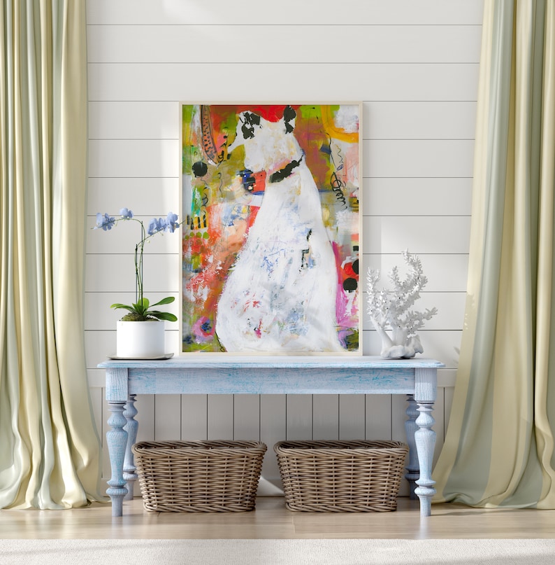 Op de afbeelding: Een lichtblauwe consoletafel met twee rieten manden eronder. Een ingelijst abstract schilderij van een witte figuur met een kleurrijke achtergrond hangt boven de tafel. Een gepotte orchidee en een wit koraalbeeld staan op de tafel.