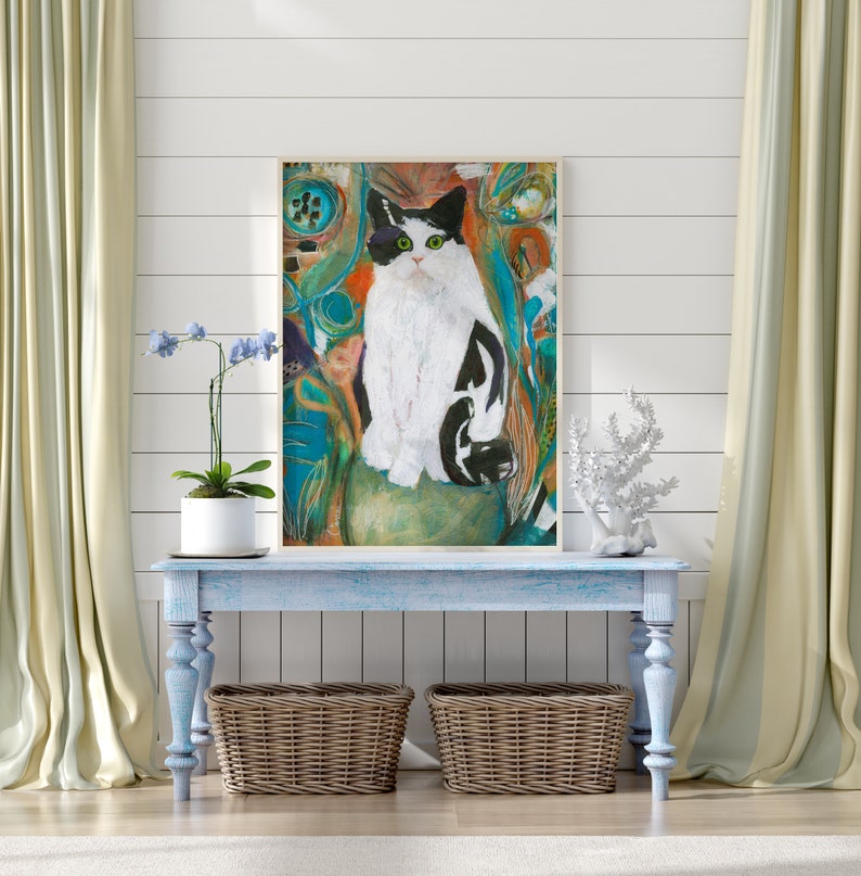 May include: A framed painting of a black and white cat sitting on a green rock. The cat is surrounded by colorful abstract shapes and lines. The painting is hanging on a white wall above a light blue console table with two wicker baskets.