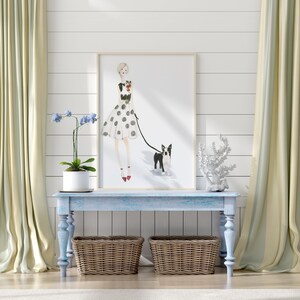 Op de afbeelding: Een ingelijste illustratie van een vrouw in een stippenjurk die een zwart-witte Boston Terrier uitlaat. De illustratie hangt boven een lichtblauwe, geverfde houten consoletafel met twee rieten manden.