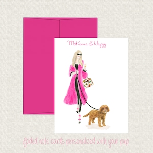 May include: A pink folded note card with a watercolor illustration of a woman in a pink coat walking a brown dog. The text "McKenna & Happy" is at the top of the card. The text "folded note cards personalized with your pup" is at the bottom of the card.