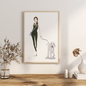 May include: Watercolour print of a woman walking a white dog. The woman wears a black outfit with a purple bag. The artwork is framed in a light wood frame and is displayed on a wooden surface.