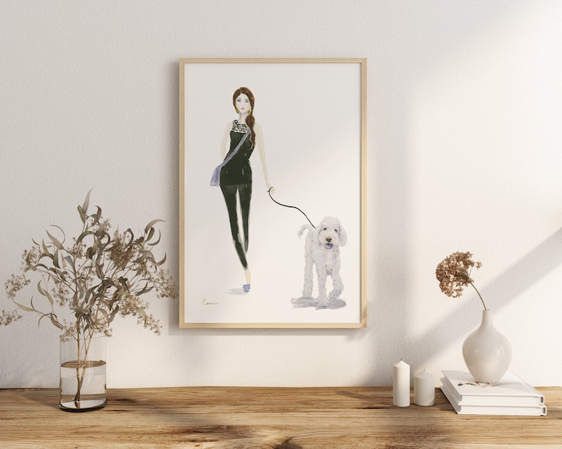 May include: Watercolour print of a woman walking a white dog. The woman wears a black outfit with a purple bag. The artwork is framed in a light wood frame and is displayed on a wooden surface.