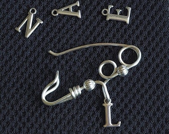 Pode incluir: Uma coleção de pingentes de letras de metal prateado, incluindo as letras B, W, R, G, E, N, A, L e U. Os pingentes estão dispostos sobre uma superfície preta texturizada.
