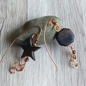 Può includere: Un paio di orecchini in filo di rame con forme di stella e ottagono in legno. Gli orecchini hanno perline marroni e sono appesi a una roccia.