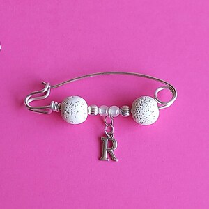 May include: A silver safety pin with white beads and a silver charm with the letter 'R' hanging from it. The pin is on a pink background.