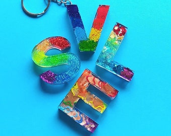 May include: A rainbow glitter keychain with the letters "S.V.L."  The letters are made of clear resin with colorful glitter and paint swirls inside.