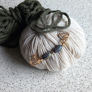 Op de afbeelding: Twee bollen garen, een groene en een witte, met een gouden veiligheidsspeld met blauwe en gouden kralen bovenop de witte bol garen.