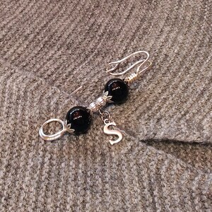 May include: A silver brooch with two black beads and a silver charm with the letter 'S' hanging from it.