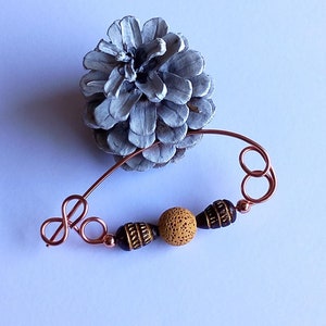 Può includere: Un fermaglio in filo di rame con un perno marrone e oro e un perno marrone e nero. Il fermaglio ha la forma di un simbolo di infinito.