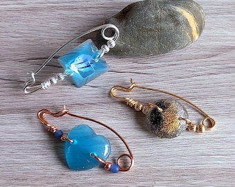 May include: Three handmade safety pin brooches with blue and gold glass accents. The brooches are made with copper and silver wire.