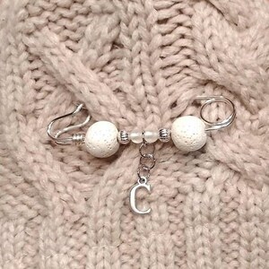 May include: A silver safety pin with white beads and a silver charm with the letter C hanging from it.