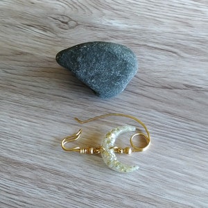 May include: A gold-toned brooch with a crescent moon design. The moon is made of clear resin with gold glitter embedded in it. The brooch has a ring attached to the top.
