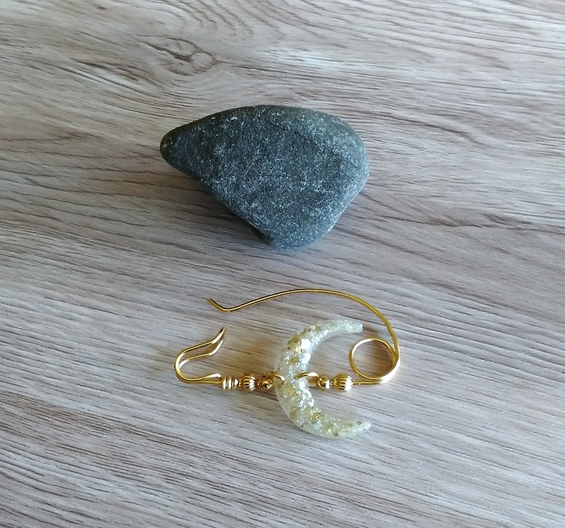 May include: A gold-toned brooch with a crescent moon design. The moon is made of clear resin with gold glitter embedded in it. The brooch has a ring attached to the top.