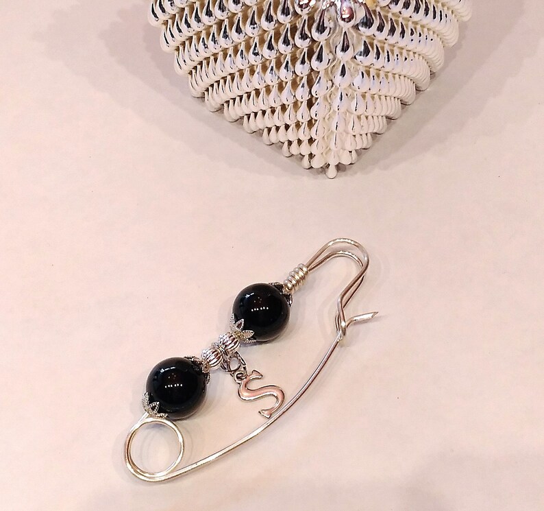 May include: A silver safety pin with two black beads and a silver charm with the letter 'S' hanging from it.