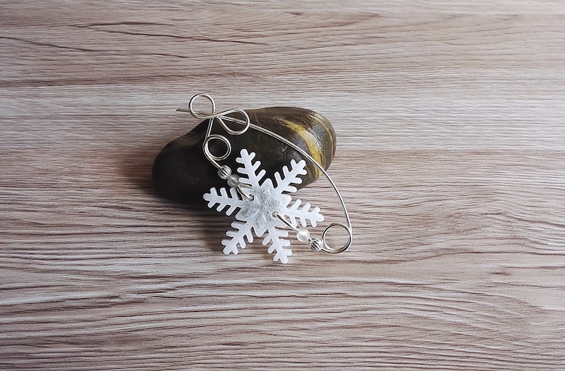 May include: A silver wire snowflake ornament with white beads hanging from it. The ornament is resting on a gray rock on a wood surface.