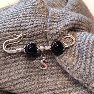 May include: A silver brooch with two black beads and a silver charm with the letter 'S'.