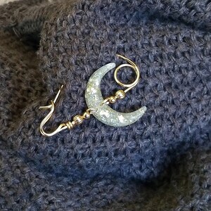 May include: A gold crescent moon brooch with glitter embedded in the resin. The brooch has a decorative loop and a pin closure.